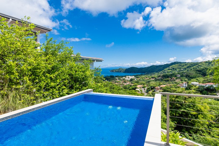 Infinity pool overlooking lush green hills and a distant blue ocean under a clear sky.