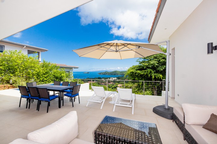 Outdoor patio with seating, umbrella, ocean view, and greenery under a blue sky.