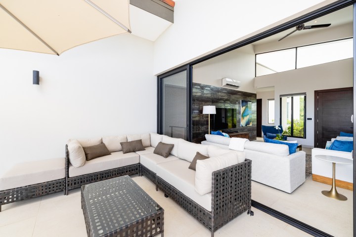 Modern patio with white sectional sofa, cushions, and table, next to interior living room with open sliding glass door.