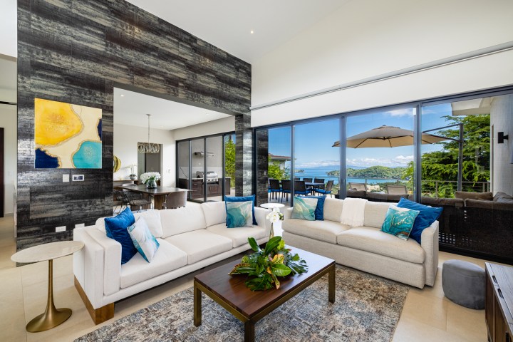 Modern living room with ocean view, white sofas, blue cushions, art, and large windows.