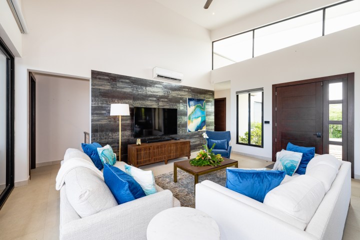 Modern living room with white furniture, blue cushions, large TV, and abstract art.