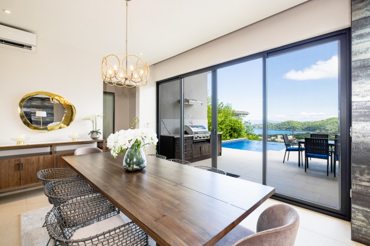 Modern dining room with large table, chandelier, and view of pool and hills through glass doors.