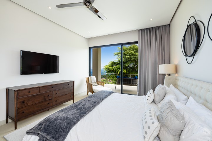 Modern bedroom with bed, dresser, TV, balcony view of trees and ocean.