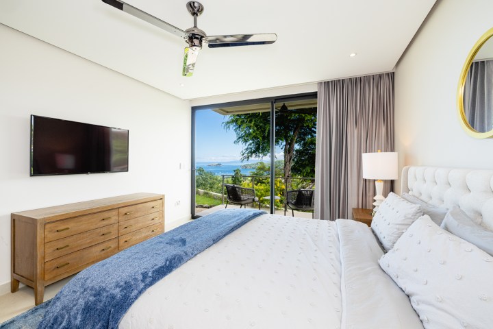 Modern bedroom with ocean view, king bed, TV, dresser, and balcony door.