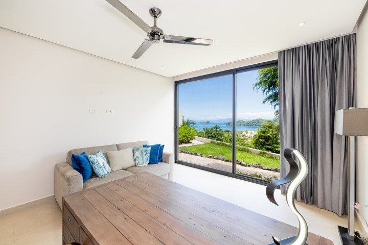Room with gray sofa, pillows, ceiling fan, and large window showing ocean view.