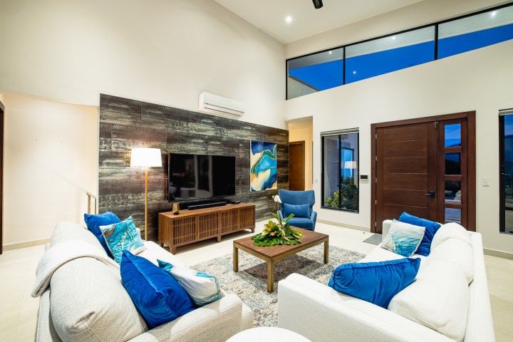 Modern living room with blue accents, large TV, and abstract artwork, featuring high ceilings and wooden elements.