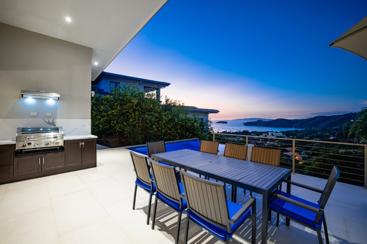 Outdoor dining area with a grill, table, and chairs, overlooking a coastal sunset view.