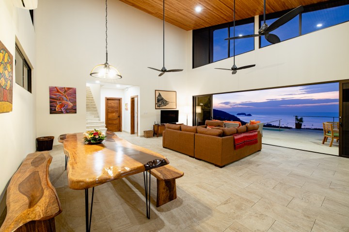 Spacious living room with wooden table, couch, and large ocean view windows at sunset.