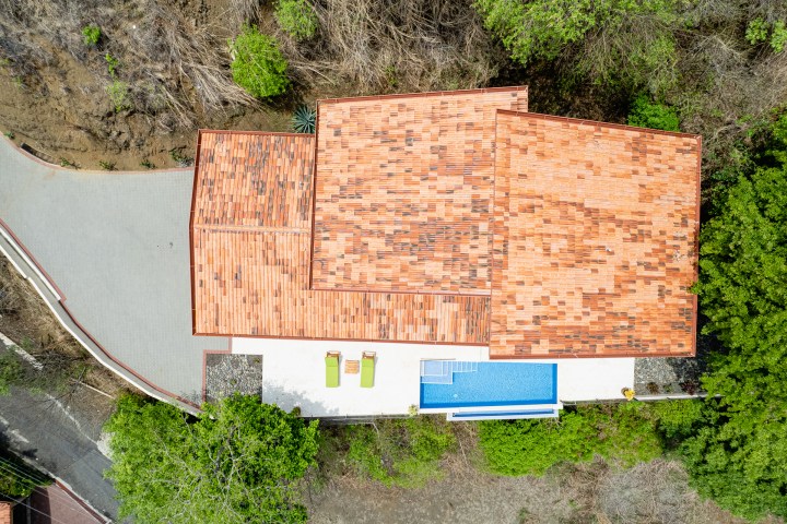 Aerial view of a house with a tiled roof, patio, and pool surrounded by trees and a driveway.