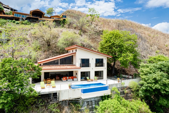 House on a hillside with a pool, trees surrounding, and clear blue sky.