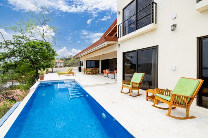 Modern house with pool, patio chairs, and trees under a blue sky with clouds.