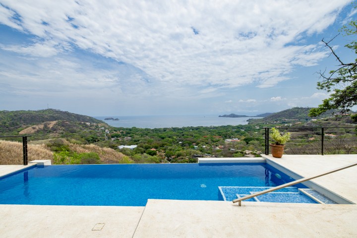 Infinity pool overlooking green hills and ocean under a partly cloudy sky.