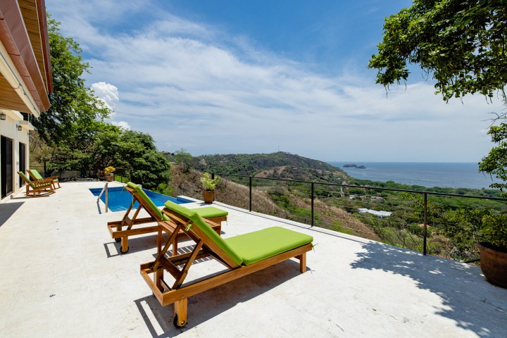Terrace with green lounge chairs, pool, ocean view, and clear sky.
