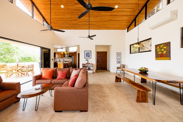 Spacious living room with brown sofas, wooden dining table, ceiling fans, and large windows.