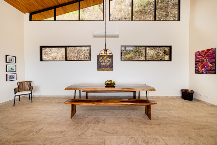 Spacious dining room with wooden table, bench, and colorful pendant light, art on walls, large windows.