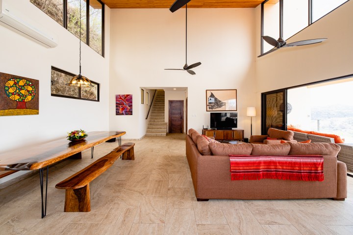 Spacious living room with high ceilings, brown sofa, wooden table, large windows, and ceiling fans.