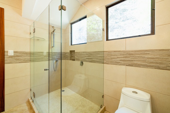 Modern bathroom with glass shower, beige tiles, two windows, and a white toilet.