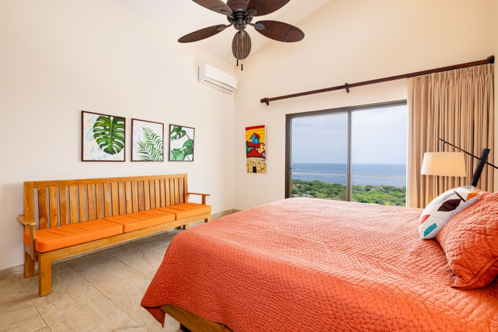 Bedroom with orange bedding, wooden bench, plant artwork, ceiling fan, and ocean view window.