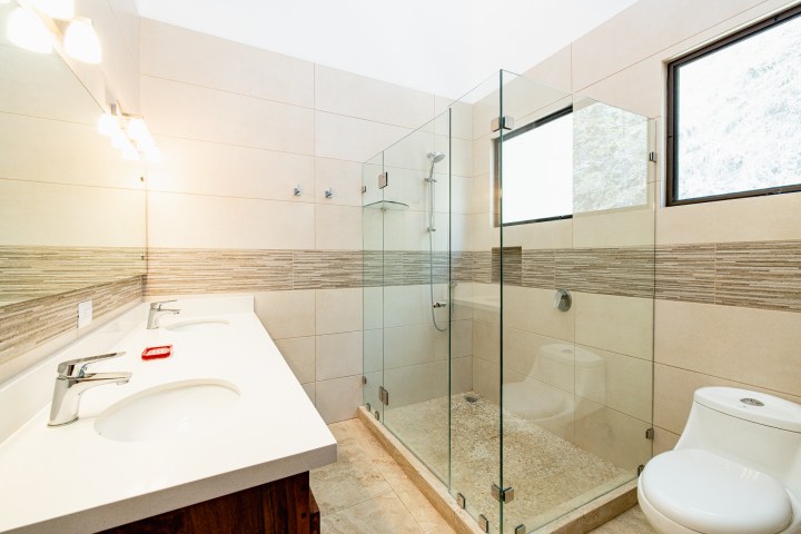 Modern bathroom with glass shower, double sink vanity, and toilet, featuring beige tiles and a window.