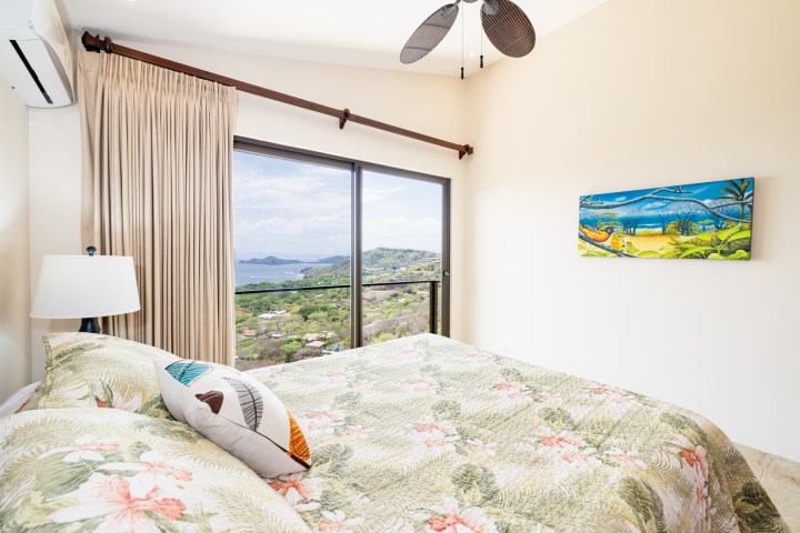 Room with floral bedding, scenic ocean view, ceiling fan, wall art, and curtain-covered windows.