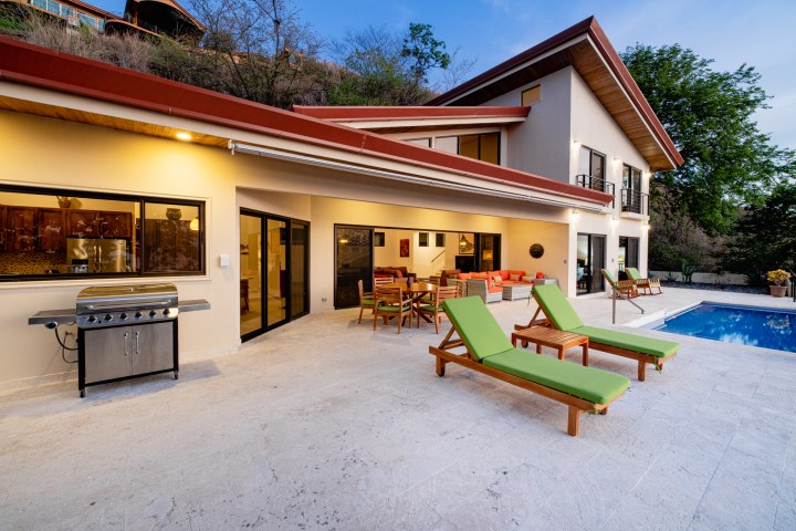 Modern house with patio, green lounge chairs, dining area, grill, and swimming pool.