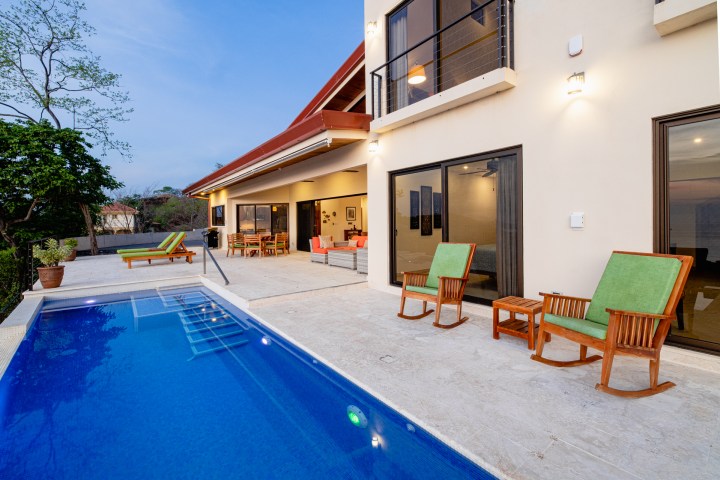 Modern patio with pool, green cushioned chairs, and a view of contemporary house.