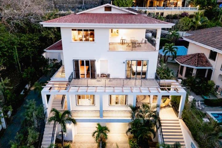 Aerial view of a modern white house with balconies, stairs, and surrounding greenery.