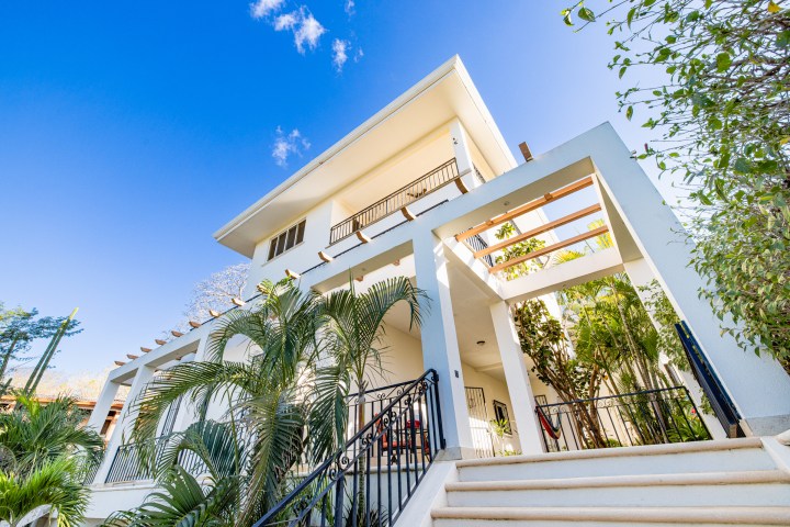 Modern white house with large windows, surrounded by green plants, under a clear blue sky.
