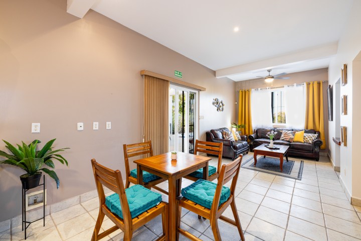Modern living room with teal-cushioned wooden dining set and brown sofas, near a window with yellow curtains.