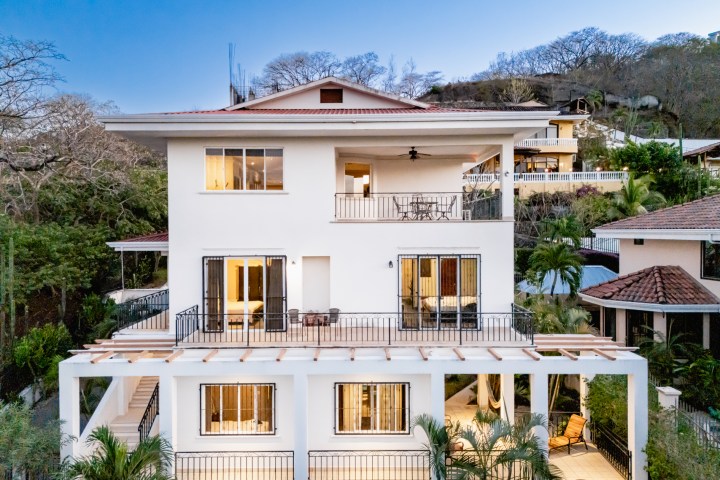 White three-story house with balconies and surrounding greenery at twilight.