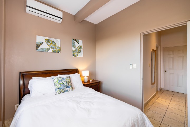 Bedroom with double bed, white sheets, wall art, nightstand, lamp, and air conditioner.