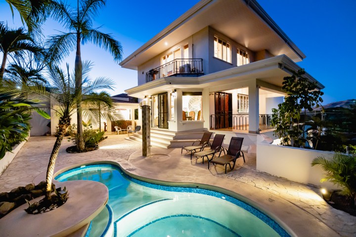 Modern two-story house with pool and palm trees at dusk.