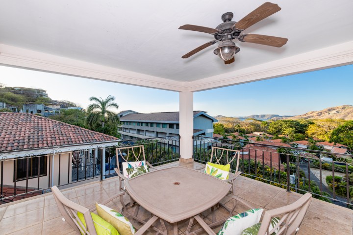 Covered patio with ceiling fan, table, and chairs overlooking houses, hills, and trees.