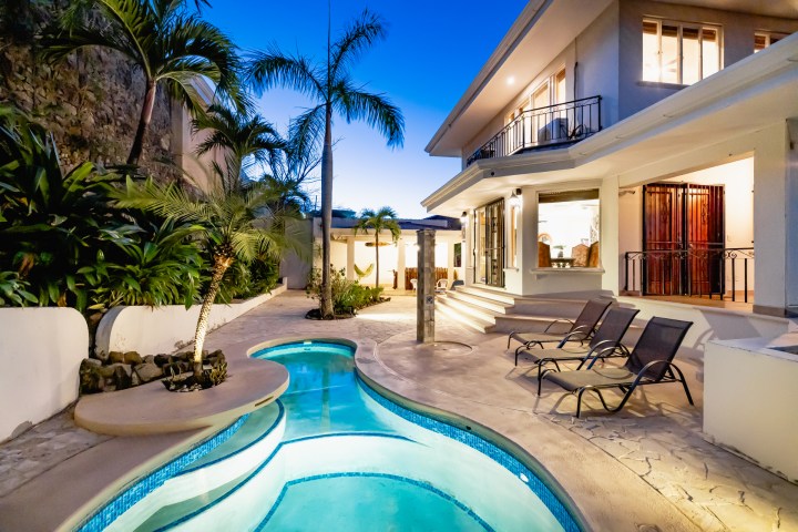 Luxurious house with pool, patio, lounge chairs, and palm trees at dusk.