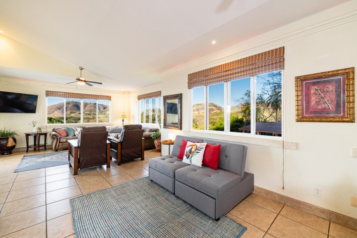 Bright living room with large windows, sofas, rugs, and a wall-mounted TV.