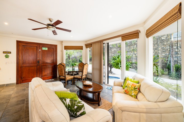Living room with beige couches, wooden table, glass doors, and tropical view.