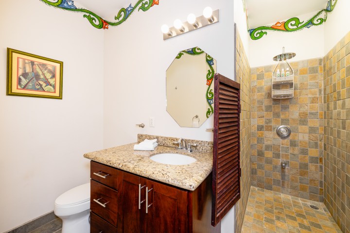 Bathroom with stone-tiled shower, wooden vanity, granite countertop, and decorative mirror.