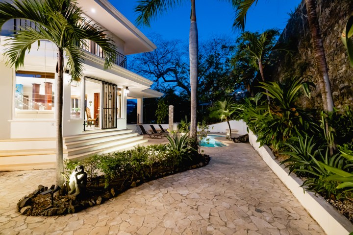 Modern villa with garden path, palm trees, and evening lighting.