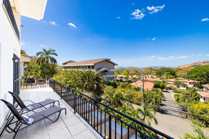 Scenic balcony view with lounge chairs overlooking tropical trees and houses under a clear blue sky.