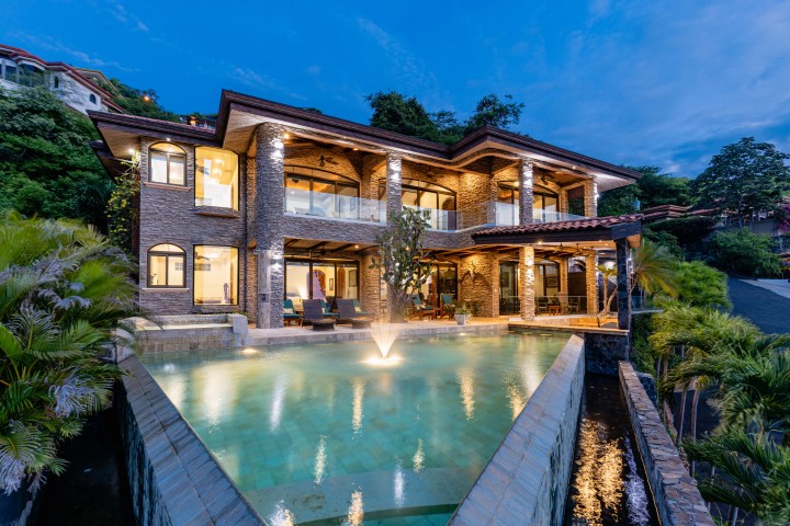 Two-story stone house with terraces and pool at dusk.