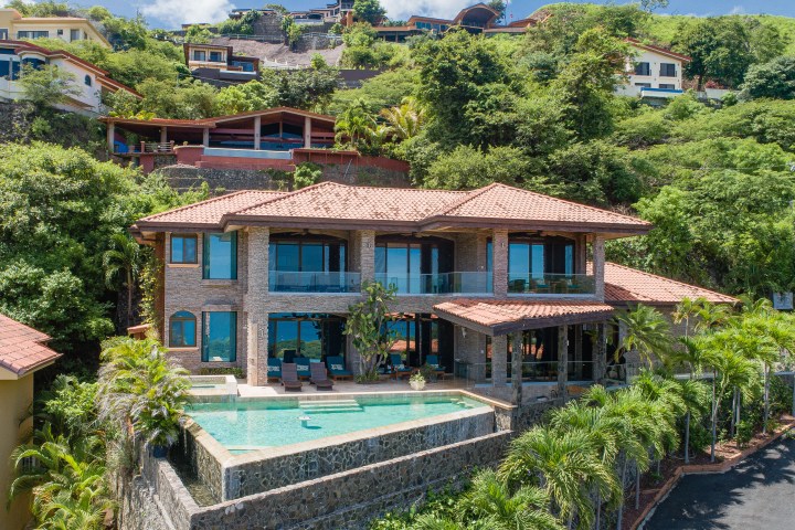 Hillside villa with pool, surrounded by lush greenery and modern architecture, under a blue sky.
