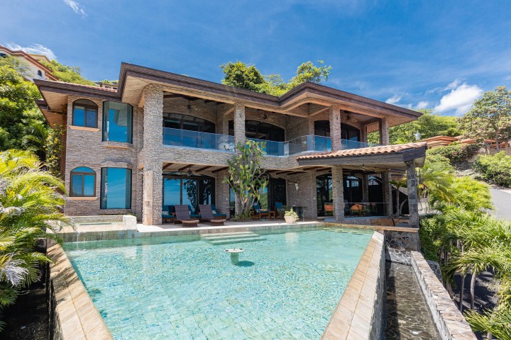 Luxury house exterior with large pool, stone walls, and tropical plants.