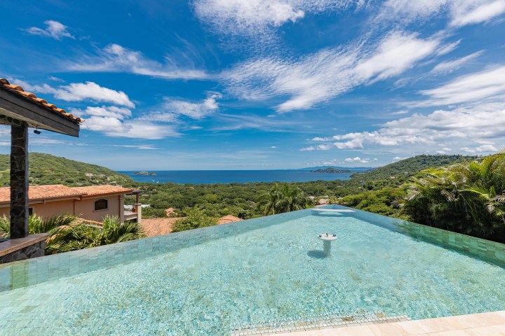 Infinity pool with ocean view, surrounded by greenery and hills under a blue sky with clouds.
