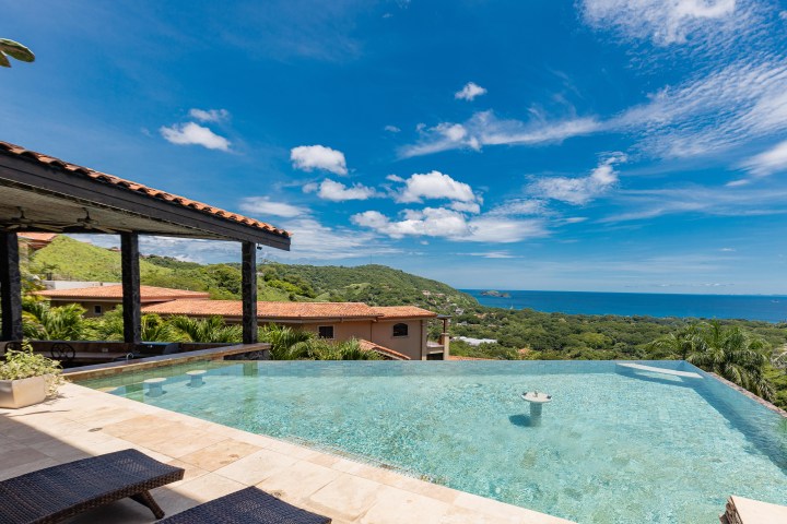 Infinity pool overlooking ocean and green hills under blue sky with clouds.