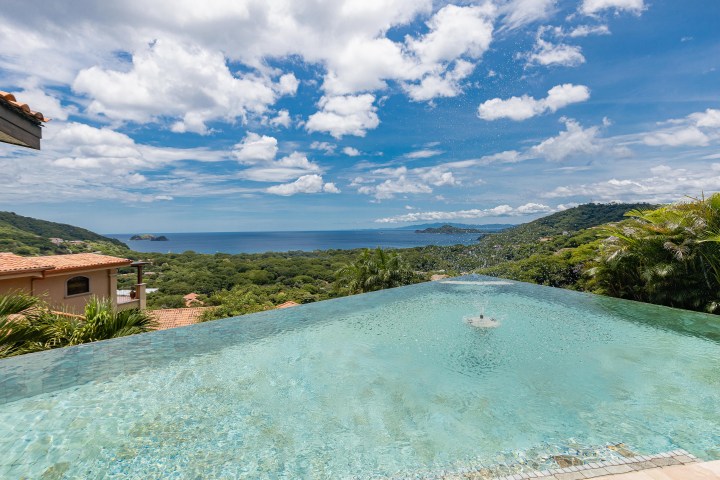Infinity pool overlooks lush hills and ocean under a blue sky with clouds.