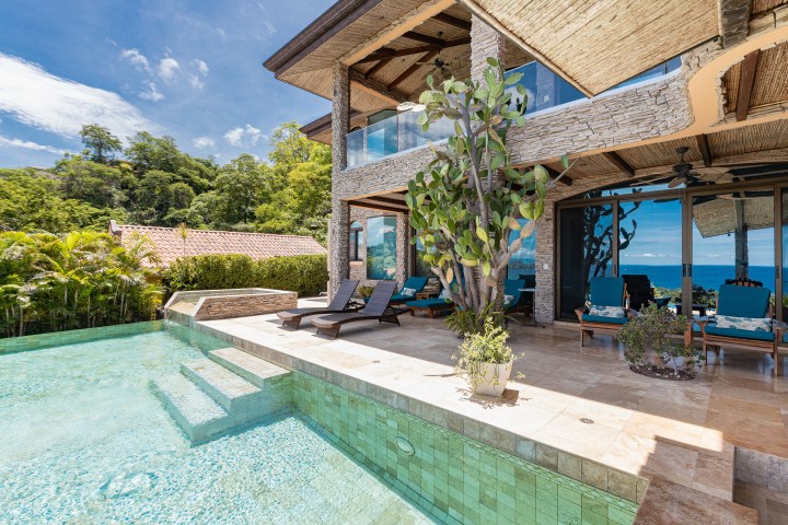 Luxury villa with pool, sun loungers, and ocean view on a sunny day, surrounded by greenery.