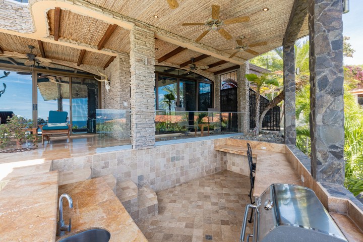 Covered outdoor patio with stone walls, kitchen area, ceiling fans, and lounge chairs beside a glass door.