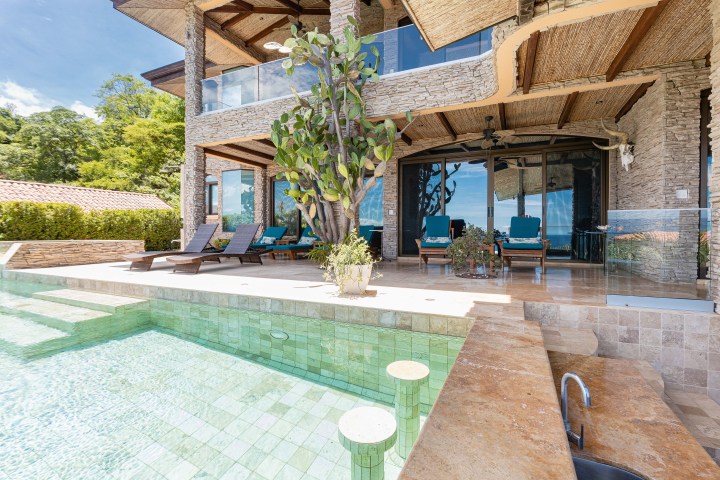 Luxurious villa patio with pool, sun loungers, and glass doors under a wooden roof.