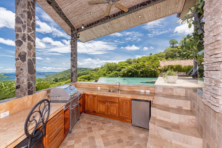 Outdoor kitchen with grill, wooden cabinets, and ocean view near infinity pool.