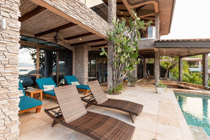 Luxury patio with wicker loungers, blue cushions, stone walls, and pool with ocean view.